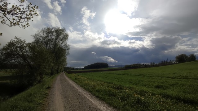 Ein Feldweg zwischen einem Bach und einem grünen Feld. Dahinter dunkle Wolken mit dramatisch durchscheinender Sonne