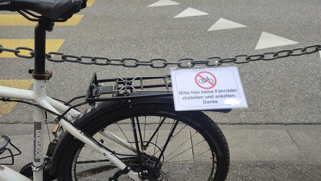 Ein Fahrrad an einer Kette geparkt. An der Kette hängt ein Schild: "Bitte hier keine Fahrräder abstellen und anketten". Das Schild wird vom Gepäcktrager des Velos darunter angehoben, weil sie so nahe sind.