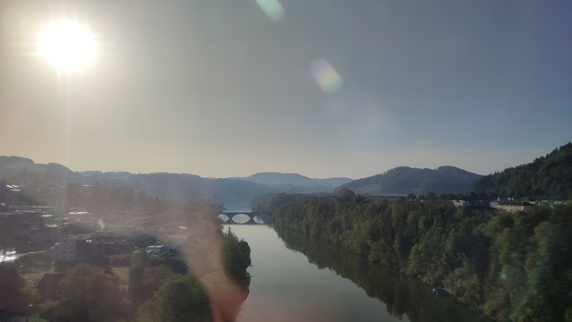 Blick von einer Brücke der Morgensonne entgegen. Darunter ein Fluss in einem grünen Tal, mit historischer Bogenbrücke im Hintergrund)