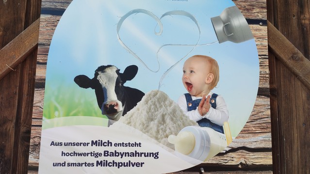 Bild eines Werbeplakats an einer Scheune: Eine Kuh und ein Baby neben einem Haufen Milchpulver; darüber eine Milchkanne, die Milch in Herzform ausleert und in diesem Haufen Milchpulver ankommt. Darunter der Text: "Aus unserer Milch entsteht hochwertige Babynahrung und smartes Milchpulver.