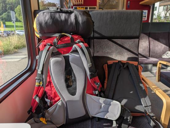 Zwei Rucksäcke, einer davon mit Schlafsack, auf zwei Sitzen eines Zugabteils.