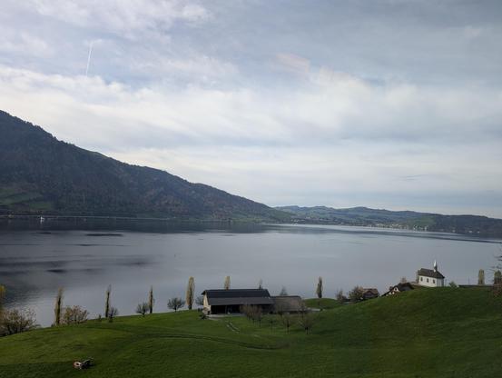 Blick über eine Wiese auf einen See mit Bergen dahinter. Rechts unden ein kleines Kirchlein.