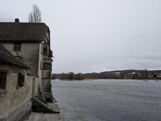 Blick über einen grauen See. Links ein historisches Gebäude, geradeaus eine bewaldete Insel und im Hintergrund eine Hügelkette. Alleine dort gibt es noch ein bisschen Schnee in Schattenhängen, sonst alles schneefrei.