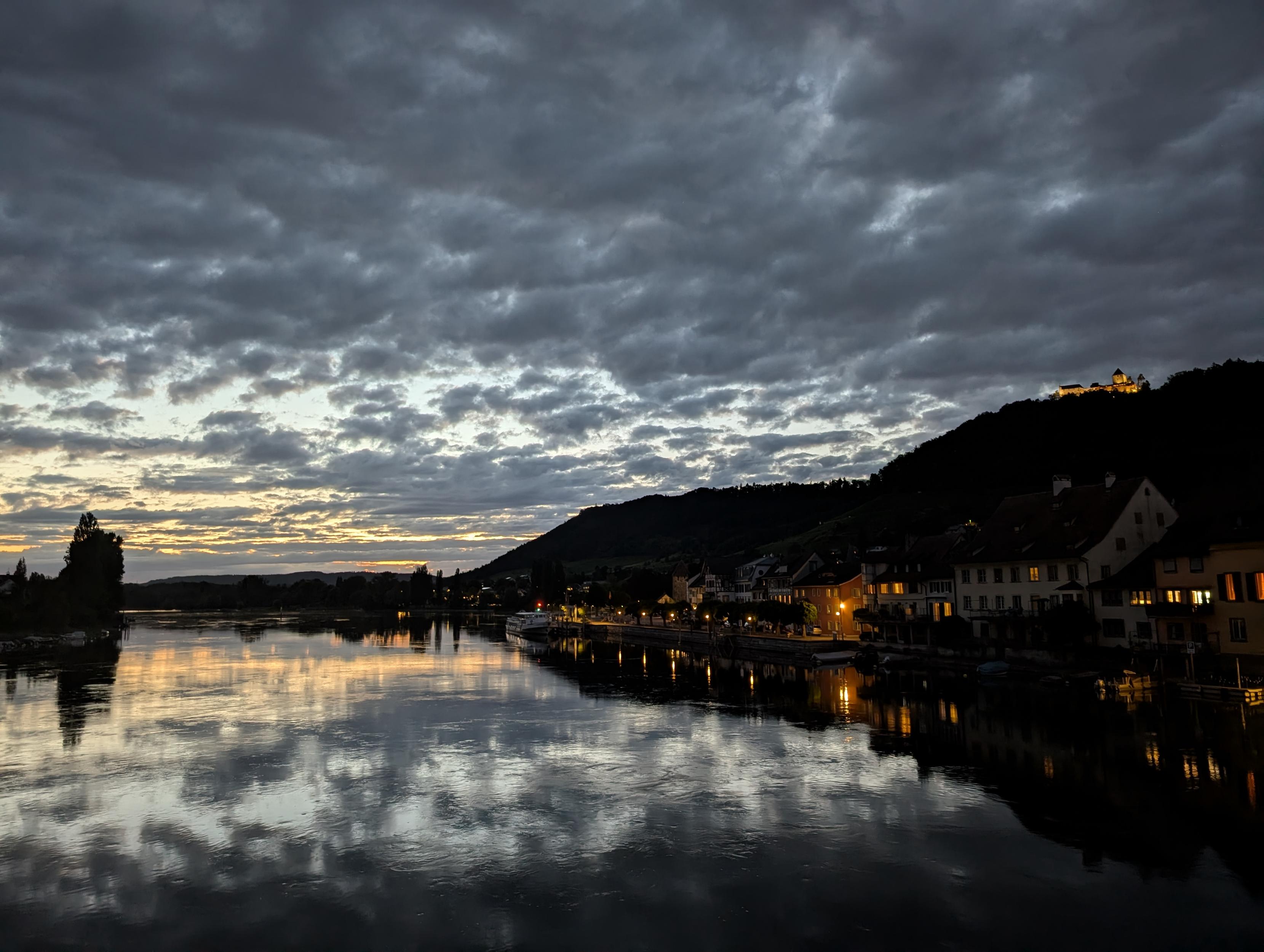 Blick von einer Brücke über eine spiegelnde Flussfläche, darüber gemusterte dunkle Wolken, von den letzten untergegangenen Sonnenstrahlen beleuchtet. Rechts das Flussufer mit historischen Häusern, darüber auf der Hügelkette thronend eine alte Burg. Fenster der Häuser und Burg sind erleuchtet.