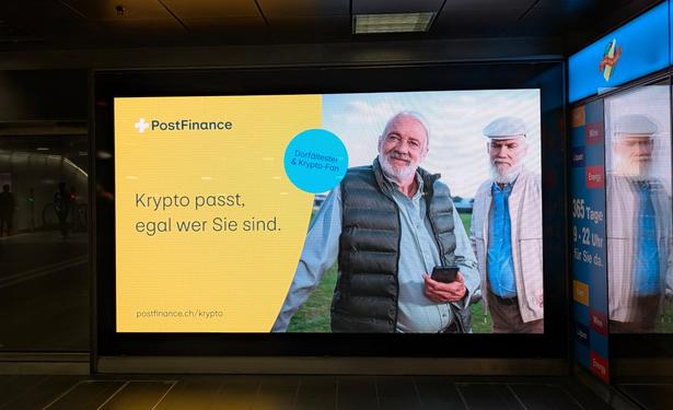 Bild eines wandgrossen Werbebildschirms mit Postfinance-Werbung: "Krypto passt, egal wer sie sind" neben zwei alten Männern, einer mit Handy und dem Label "Dorfältester & Krypto-Fan"