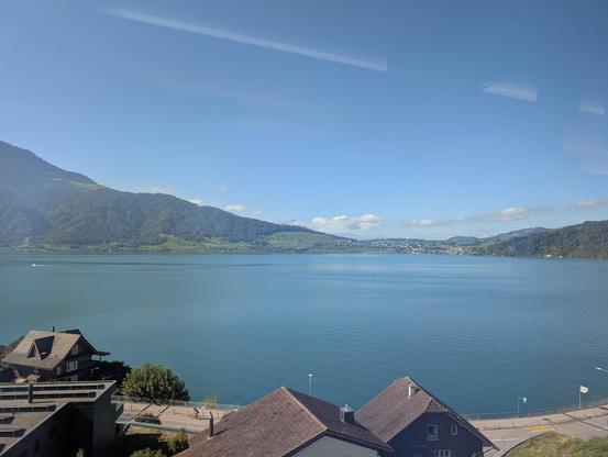 Blick über ein paar Dächer und einen blauen See, umgeben von Bergen unter einem blauen Himmel. Ein paar Reflexionen und Unschärfen bedingt durch das Zugfenster.